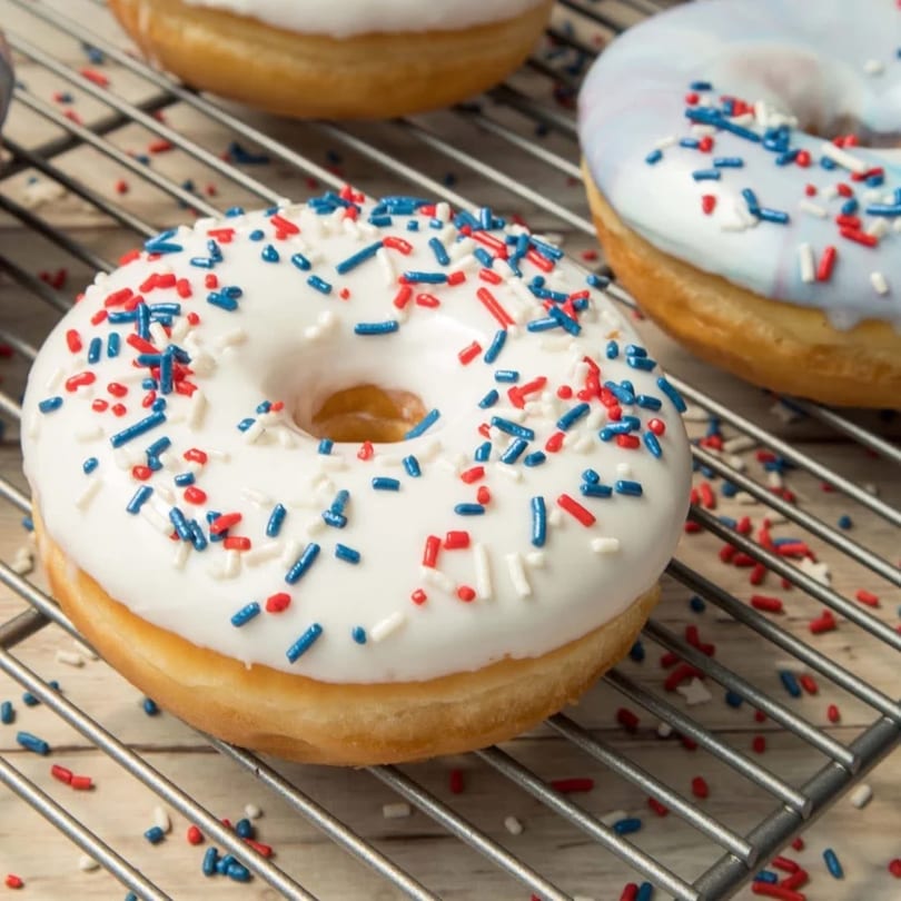 White and blue frosted donuts with red, white, and blue sprinkles on a cooling rack.