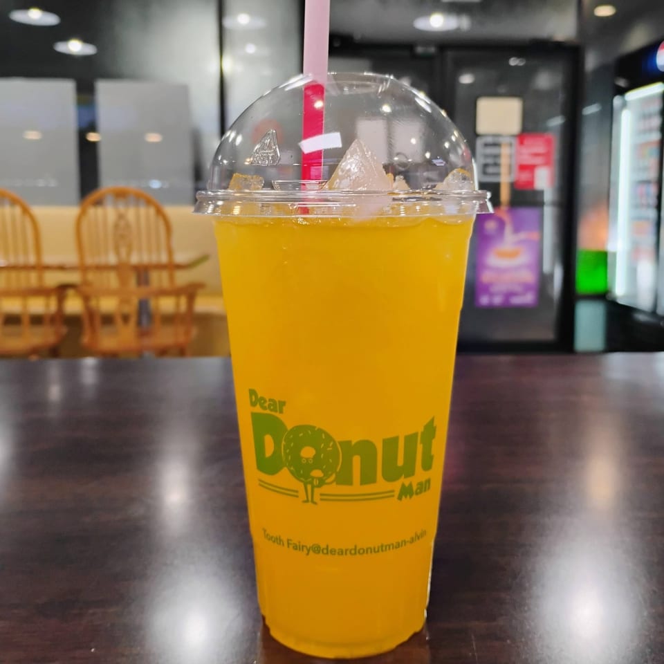 A yellow drink with ice and a pink straw in a "Dear Donut Man" cup on a dark table.