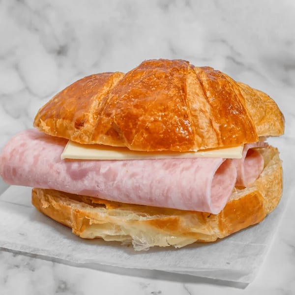A golden-brown croissant sandwich filled with thinly sliced ham and a slice of white cheese.