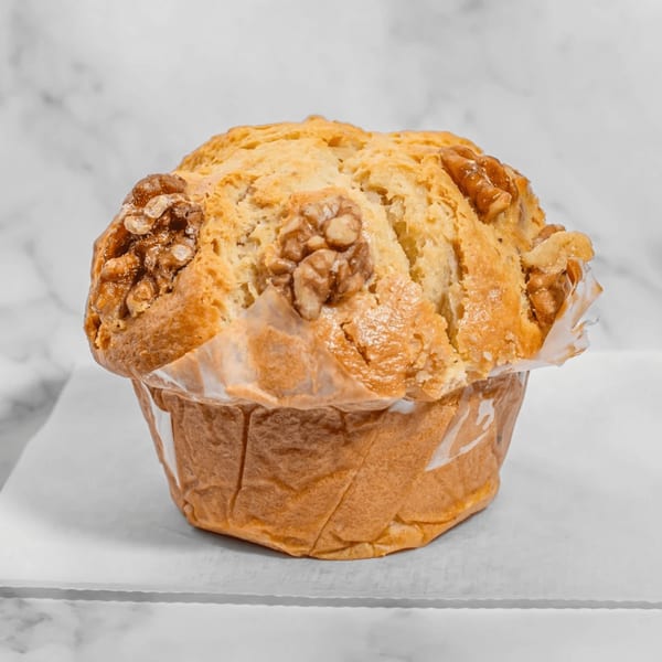 Golden-brown muffin with walnuts on top, in a paper liner, against a white marble background.
