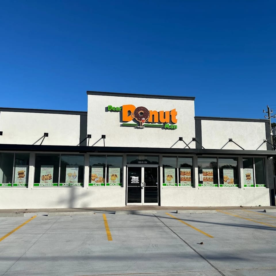 The exterior of Dear Donut Man, a white building with a large orange and green sign, under a clear blue sky.