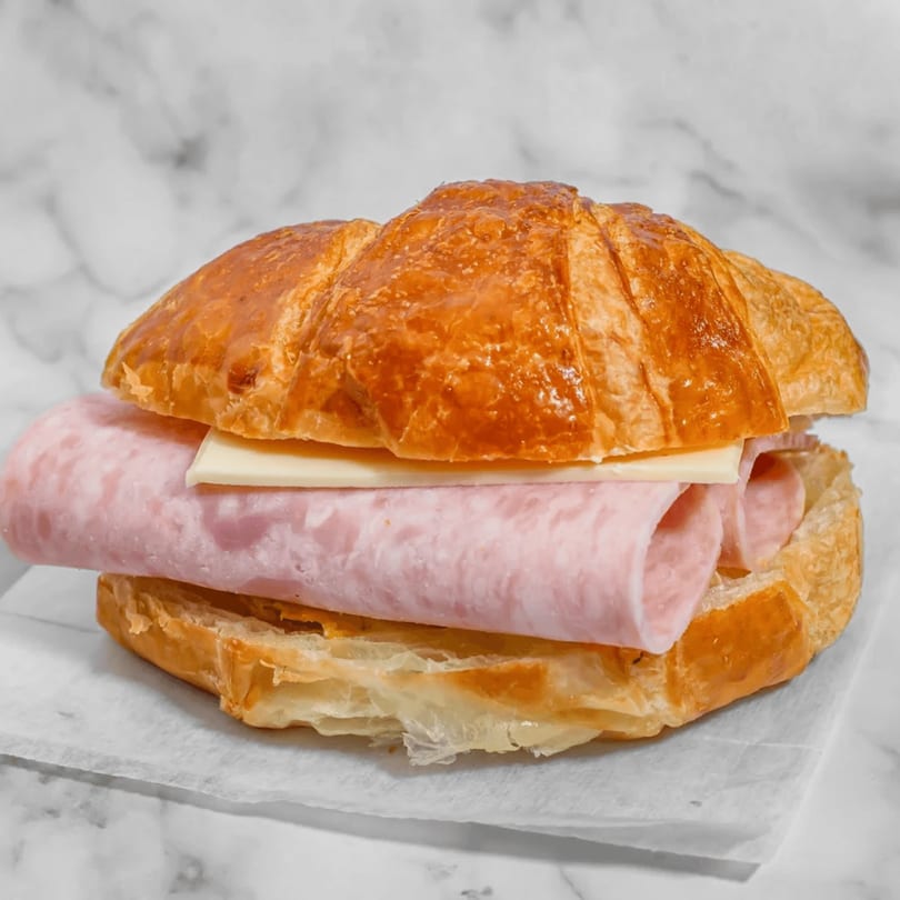 Golden-brown croissant sandwich filled with thinly sliced ham and a slice of white cheese.